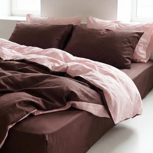 May include: A bed with a dark brown fitted sheet, duvet cover, and pillowcases. The duvet cover and pillowcases are reversible, with a light pink color on the reverse side. The bed is in a bright room with two windows.
