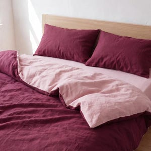 May include: A bedding set featuring a deep burgundy duvet cover and two matching pillowcases, paired with a light pink flat sheet. The duvet cover is folded back to reveal a contrasting light pink underside, creating a layered colour effect.