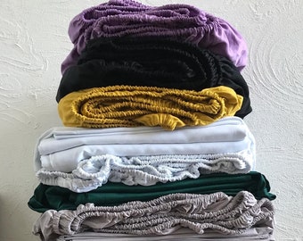 May include: A stack of 11 fitted sheets in various colors, including purple, black, yellow, white, green, gray, and beige. The sheets are folded and stacked on top of each other on a black table.