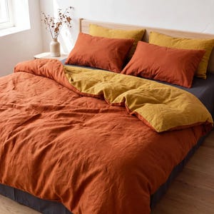 May include: A bedding set featuring a reversible duvet cover in burnt orange and mustard yellow, paired with two matching burnt orange pillowcases and two mustard yellow pillowcases, all resting on a dark gray fitted sheet on a wooden bed frame.