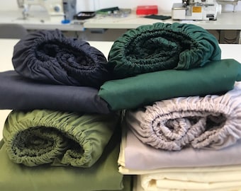 Puede incluir: Una pila de cuatro sábanas bajeras de diferentes colores: azul marino, verde bosque, verde oliva y gris claro. Las sábanas están enrolladas y apiladas unas sobre otras.