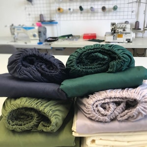 Puede incluir: Una pila de cuatro sábanas bajeras de diferentes colores: azul marino, verde bosque, verde oliva y gris claro. Las sábanas están enrolladas y apiladas unas sobre otras.