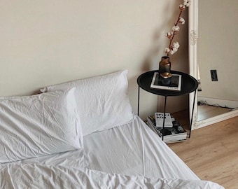 Puede incluir: Una escena de dormitorio minimalista con una cama con sábanas y almohadas blancas. Una pequeña mesa auxiliar negra sostiene un jarrón con tallos de algodón y un libro. Un gran espejo refleja la habitación, realzando la estética brillante y aireada.