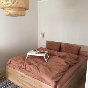 May include: A bedroom scene featuring a wooden bed with terracotta-colored bedding and pillows. A white tray with breakfast items sits on the bed. A woven pendant light hangs above the bed, and a patterned rug is at the foot of the bed.
