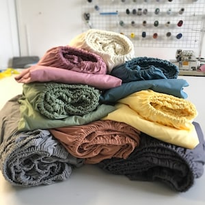Puede incluir: Una pila de ocho fundas de sábanas ajustables en varios colores, incluyendo gris, beige, verde, amarillo, azul y rosa. Las sábanas están enrolladas y apiladas sobre una superficie blanca.