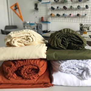 Puede incluir: Cuatro sábanas bajeras de diferentes colores, incluyendo blanco, beige, marrón y verde, están apiladas sobre una mesa blanca. Las sábanas están dobladas y enrolladas, mostrando la textura de la tela.