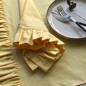 Peut inclure: Un napperon jaune à volants avec une serviette jaune à volants pliée dessus. Une assiette blanche avec une fourchette et un couteau est sur le napperon.