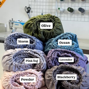 May include: A stack of fitted bed sheets in various colors, including olive, storm, ocean, pink fog, lavender, powder, and blackberry. Each sheet is labeled with its color name. The sheets are neatly folded and displayed.