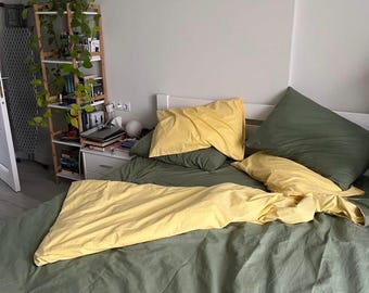 Puede incluir: Una cama con ropa de cama verde oliva y almohadas amarillas y una manta amarilla. A la izquierda de la cama hay una estantería de madera con plantas y libros. La habitación tiene una paleta de colores neutros.