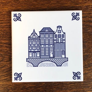 May include: A white ceramic tile with a blue and white illustration of three buildings with a bridge in front of them. The buildings have multiple windows and a decorative design. The tile has a blue fleur-de-lis design in each corner.