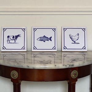 Może przedstawiać: Trzy kwadratowe białe płytki ceramiczne z ciemnoniebieskimi ilustracjami. Każda płytka ma ciemnoniebieską obwódkę z ozdobnymi akcentami w rogach. Lewa płytka przedstawia krowę, środkowa rybę, a prawa płytka kurę.
