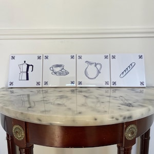 May include: Four white ceramic tiles with blue line drawings of a coffee maker, coffee cup with croissant, pitcher, and baguette. Each tile has a blue decorative corner design. The tiles are displayed on a marble-topped table.