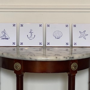 Op de afbeelding: Vier witte vierkante tegels met blauwe nautische ontwerpen: een zeilboot, een anker, een zeeschelp en een zeester. Elke tegel heeft een decoratief blauw hoekontwerp. De tegels worden getoond op een tafel met marmeren blad.