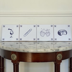 Puede incluir: Cuatro baldosas cuadradas blancas con ilustraciones azules de comida y bebida. Las imágenes incluyen una copa de vino con vino vertiéndose, una baguette, dos jarras de cerveza y una rueda de queso. Cada baldosa tiene un borde decorativo azul.