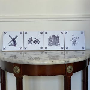 Puede incluir: Cuatro baldosas de cerámica blancas con ilustraciones azules. Las baldosas representan un molino de viento, una bicicleta, edificios y un diseño floral. Cada baldosa tiene un borde azul decorativo. Las baldosas se exhiben sobre una mesa con tapa de mármol.