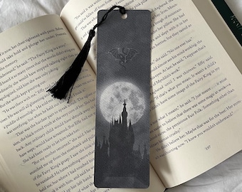 May include: A black and white bookmark featuring a silhouette of a castle with a full moon and a bat in the sky. The bookmark has a black tassel.