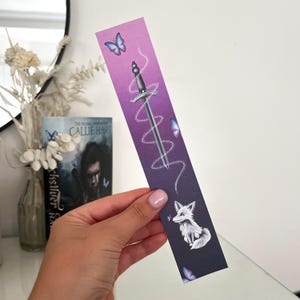 May include: A bookmark with a purple gradient background, depicting a silver sword, a white fox, and blue butterflies. The bookmark has a fantasy theme, with a sword design and a fox illustration. The bookmark is held by a person.