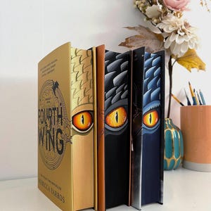 Op de afbeelding: Een stapel fantasyboeken met drakenoogontwerpen. Het boek aan de linkerkant is goudkleurig met de titel "Fourth Wing" van Rebecca Yarros. De andere boeken hebben zwarte, bruine en blauwe ruggen met drakenoogillustraties.