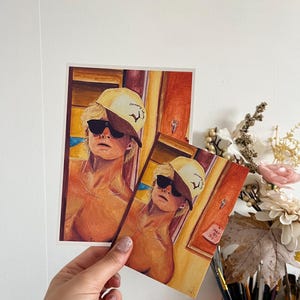 Op de afbeelding: Twee rechthoekige kunstdrukken met een aquarelillustratie van een persoon met blond haar die een geel-bruine honkbalpet en een zwarte zonnebril draagt. De persoon is afgebeeld in een warm getint interieur met een dunne zilveren ketting om de nek.