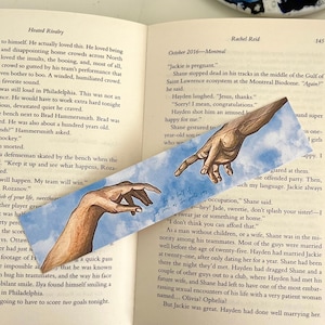 May include: A rectangular paper bookmark featuring a printed illustration of two hands reaching toward each other against a light blue sky with white clouds. The bookmark is placed diagonally across the open pages of a printed book with text.