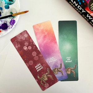 May include: Three rectangular bookmarks with gradient backgrounds. One is burgundy with the text "AMAZE AMAZE AMAZE!" and a creature illustration. Another is pink and purple with "WHY STUPID, QUESTION?" and a creature. The last is green with "FIST MY BUMP!" and a creature.