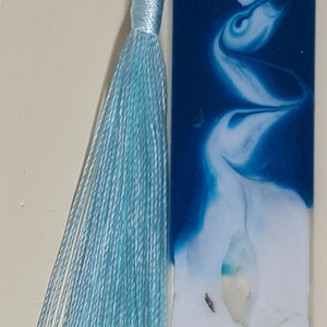 May include: A blue and white resin bookmark with a white seashell and a starfish on a sandy beach background. The bookmark has a light blue tassel.