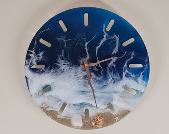May include: A blue and white resin clock featuring a beach scene. The clock face has a white wave design with a brown wooden hand and gold accents. The clock face also includes a starfish and seashells.