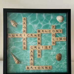 May include: A framed artwork featuring a turquoise blue background with a white water ripple pattern. The artwork includes a scrabble tile crossword puzzle with the words "HURRICANE", "BALCONY", "CRUISE", "LIMO", "BAYONNE", and "P.E.S.". The artwork also includes three seashells.