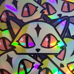 May include: Holographic cat stickers with a stylized design. The stickers feature a cat face with large, orange eyes, a black nose, and a diamond shape on the forehead. The stickers have a rainbow sheen.