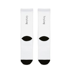 May include: A pair of white socks with black toe and heel. The left sock says "Trying" and the right sock says "Crying".