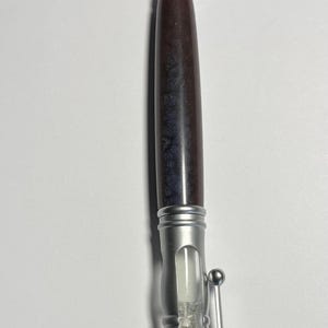 Puede incluir: Un bolígrafo plateado con un barril de madera oscura y un reloj de arena en el medio. El reloj de arena está lleno de arena blanca.