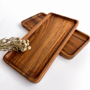 May include: Three rectangular wooden trays with rounded corners are displayed. The trays are a warm brown color, showcasing the natural wood grain. A sprig of dried flowers with small, round, beige buds rests near the trays, adding a touch of natural decor.