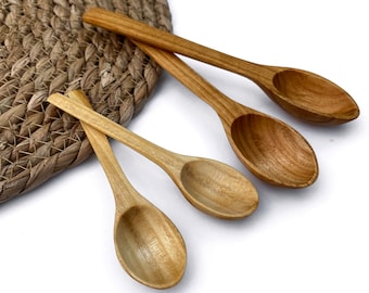 Könnte beinhalten: Vier kleine Holzlöffel mit ovalen Schalen und langen Griffen sind auf einer weißen Oberfläche angeordnet. Die Löffel variieren in der Farbe von hell- bis mittelbraun. Ein gewebter Platzdeckchen ist teilweise im Hintergrund zu sehen.