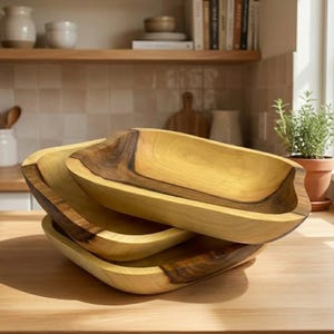 Könnte beinhalten: Ein Stapel von drei Holzservierschalen. Die Schalen sind rechteckig mit abgerundeten Ecken und einer natürlichen Holzmaserung. Die obere Schale hat einen dunkelbraunen Rand, während die anderen heller sind. Die Schalen stehen auf einer Holzoberfläche.