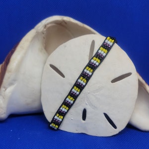 May include: A beaded bracelet with black, white, yellow, and purple beads. The bracelet is lying on a white seashell.