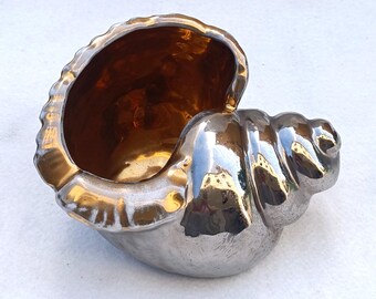 Puede incluir: Un cuenco decorativo con forma de concha, con exterior plateado e interior dorado. El cuenco tiene una superficie texturizada y un acabado brillante, que refleja la luz. La abertura es ancha y el diseño general recuerda a una concha de nautilus.