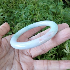 May include: A jade bangle bracelet in shades of pale green and white, with subtle pinkish-red inclusions. The smooth, polished surface reflects light, highlighting the natural variations in the stone. The bracelet is held in an open hand against a backdrop of green foliage.