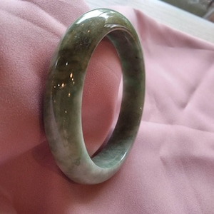 以下が含まれることがあります： 丸みを帯びた形状の滑らかで光沢のある翡翠のバングルブレスレット。石の表面全体に、落ち着いたセージグリーン、深いフォレストグリーン、半透明のオフホワイトの色調が渦巻くような模様を描いており、変化に富んだ外観を呈しています。