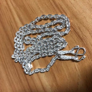 Könnte beinhalten: Eine silberne Kettenkette mit einem detaillierten, komplizierten Design. Die Kette besteht aus kleinen, miteinander verbundenen Quadraten und hat einen Verschluss mit einem dekorativen Design.