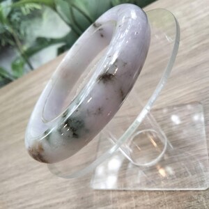 May include: A smooth, polished bangle bracelet crafted from light lavender stone with natural dark green and black mineral inclusions throughout the surface. The circular bangle features a rounded profile and a glossy finish, displaying unique patterns.