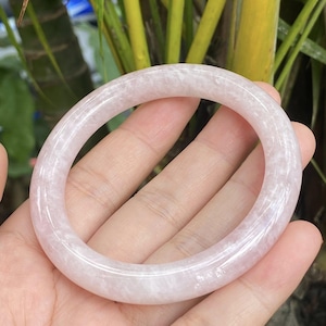 May include: A light pink jade bangle bracelet. The bracelet is a solid, circular band with a smooth, polished surface. The color is a soft, translucent pink with subtle variations in tone. The bangle is held in a hand against a background of green foliage.