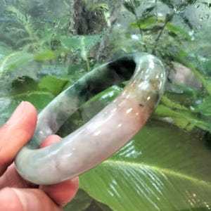 May include: A smooth, polished bangle bracelet crafted from natural stone, featuring a gradient of colours ranging from translucent white and soft peach to deep forest green with dark, mottled inclusions throughout its rounded, circular form.