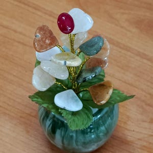 May include: A small green ceramic vase with a bouquet of heart-shaped stones in various colors, including white, brown, green, and red. The stones are attached to gold wire stems.