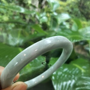 May include: A smooth, polished, light gray jade bangle bracelet with a rounded profile and a translucent, mottled appearance. The circular band features subtle variations in tone and a glossy finish that reflects light across its curved surface.