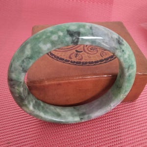 May include: A jade bangle bracelet with a marbled appearance. The bracelet features shades of green and white, with darker green inclusions. The smooth, polished surface reflects light, highlighting the natural variations in the stone. This piece is a classic jewelry item.