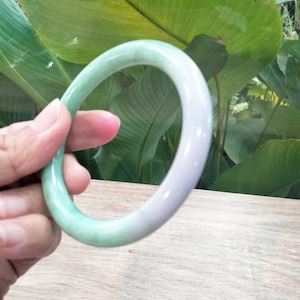 Könnte beinhalten: Ein Jade-Armreif mit glatter, polierter Oberfläche. Das Armband geht von einem hellen Grün in eine blasslila Farbe über. Der Armreif wird vor dem Hintergrund von grünem Laub und einer Holzoberfläche gehalten.