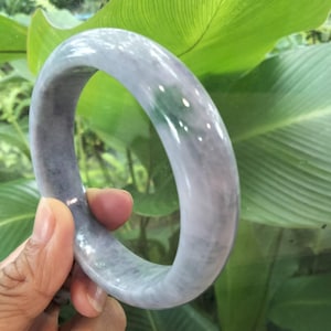May include: A smooth, polished bangle bracelet crafted from translucent lavender and grey stone. The circular band features a rounded profile with subtle variations in color, displaying a cloudy, marbled texture throughout the polished surface.
