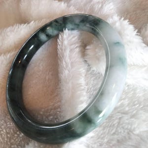 May include: A smooth, polished bangle bracelet crafted from natural stone, featuring a distinct color gradient that transitions from a deep, mottled forest green on one side to a translucent, milky white on the opposite side.