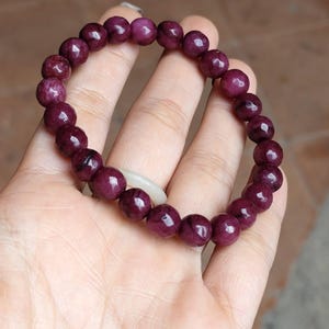 Könnte beinhalten: Ein Armband aus tief burgunderfarbenen Perlen mit poliertem Finish. Die runden Perlen sind zu einem Kreisdesign aufgefädelt. Ein weißes, ovales Objekt ist in der Mitte des Armbands sichtbar. Das Armband wird in einer Hand gehalten.