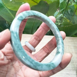 May include: A jadeite jade bangle bracelet and a jade ring. The bracelet is a wide band of pale green and white jade. The ring is a thin band of white jade. The jewelry is held in a hand.